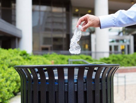 A person's hand, wearing a light blue long-sleeved shirt, is seen dropping a crumpled clear plastic bottle into a black, cylindrical rubbish bin with vertical slats, situated outdoors on a paved surface. The bin is positioned against a backdrop of green bushes and modern office or commercial buildings with large windows and structural columns. The scene appears to be in a public or commercial area, possibly near a business premises in Crawley, Sussex, as suggested by the context of house clearance and rubbish removal services. Natural daylight illuminates the scene, highlighting the texture of the plastic bottle and the metallic finish of the bin. The focus is on the act of discarding the plastic waste responsibly, aligning with environmental and waste management practices associated with local rubbish removal companies like House Clearance Crawley.