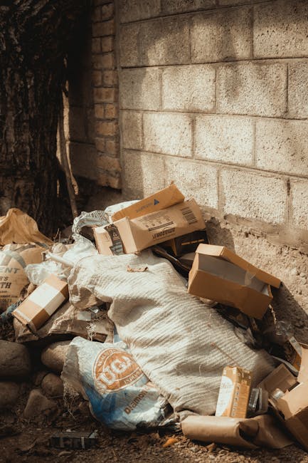 A pile of black rubbish bags, some torn open, is stacked in front of a red metal door on an outdoor building wall. The door features graffiti tags in white and pink spray paint, and a small white sign