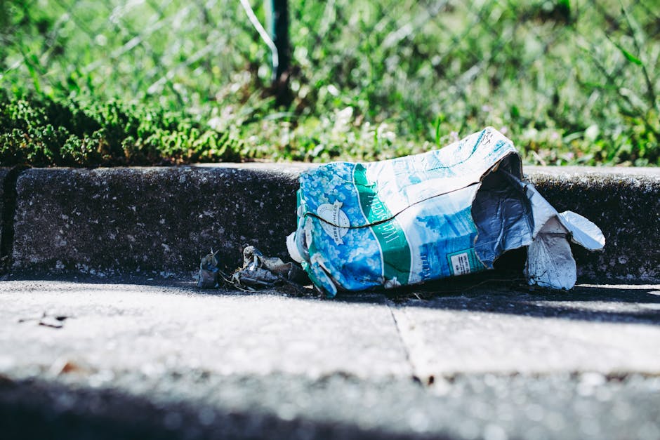 A narrow, cluttered alleyway between two brick and corrugated metal walls, both covered in multicoloured graffiti. The ground is uneven, composed of dirt, patches of moss, and scattered debris. Variou