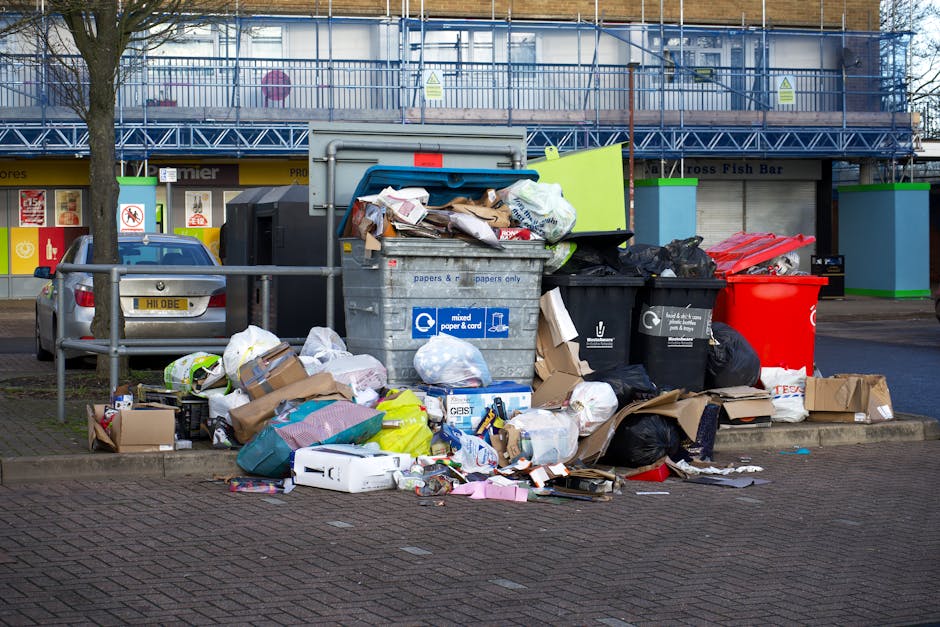 A collection of overflowing waste bins and scattered rubbish on a paved pavement in an urban area, with a silver car parked nearby and a blue construction scaffold covering a building in the backgroun