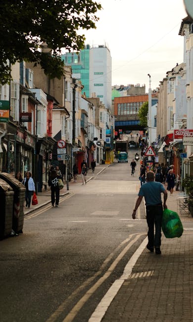 A collection of overflowing waste bins and scattered rubbish on a paved pavement in an urban area, with a silver car parked nearby and a blue construction scaffold covering a building in the backgroun